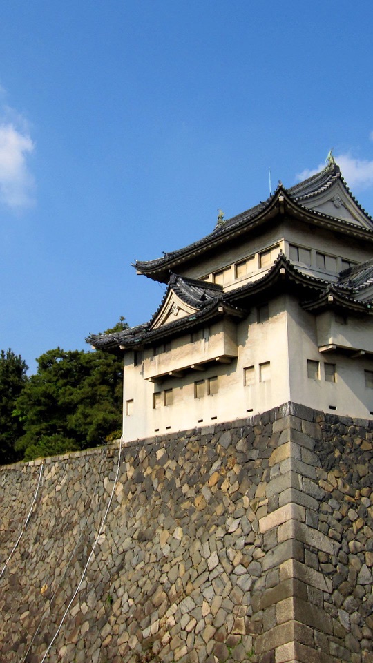 青空を背に白壁と黒瓦の櫓が石垣の上にそびえ左手に緑の木立が見えます。