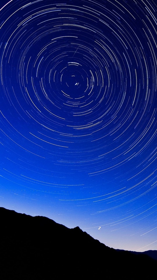 夜空に星が同心円を描く軌跡と、丘の黒い稜線を写した縦長の写真です。