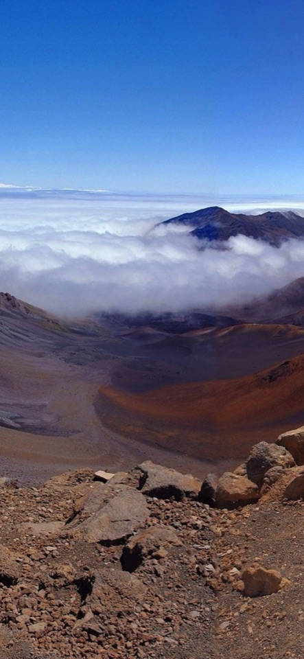 青空の下、褐色の火山礫に覆われた広大な火口が広がり、雲海が稜線に沿ってたなびく高山の写真。