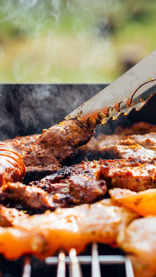 煙の立つ炭火のグリルで、こんがり焼ける肉をトングでつかむクローズアップ写真。