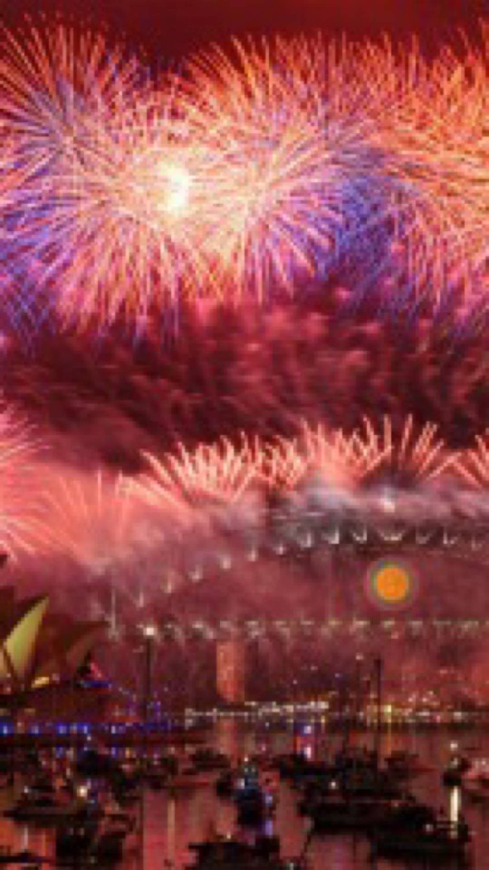 夜の港の空に、赤や紫、金の大輪の花火が幾重にも開き、橋や船の明かりが連なる縦長の写真。
