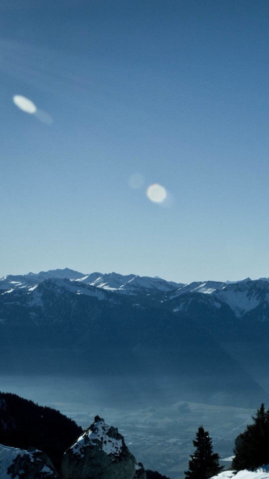青空の下、霞のかかった雪山の連なりと、麓の谷を見下ろす高所からの眺めです。