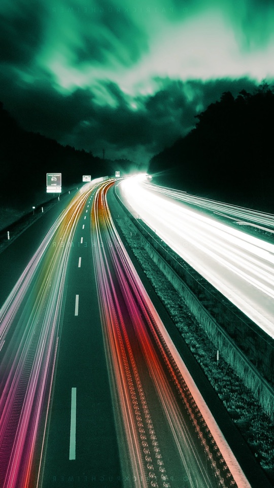 緑がかった薄暮の空の下、高速道路を走る車のライトが色の光跡となって伸びる写真。
