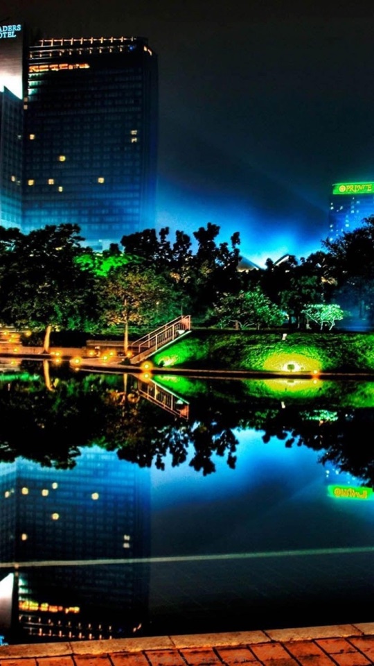 夜、高層ビルのそばでライトアップされた緑の庭園と橋が、水面に鮮やかに映り込む夜景写真。