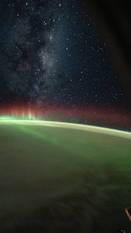 国際宇宙ステーションから見た夜の地球。緑のオーロラが地球の縁に沿って弧を描き、その上に赤い光と天の川の星が広がる。