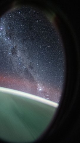 宇宙船の丸い窓越しに見た天の川。窓枠の暗い縁の内側に星の帯が広がり、下に地球の縁の光が見える。