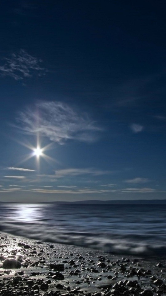 夜の空に月が輝き、その光が穏やかな海と小石の浜を照らす静かな写真。