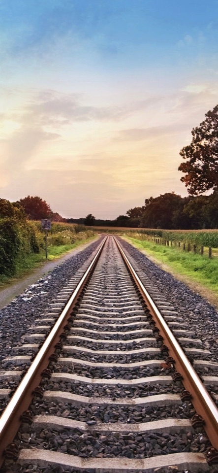 淡い夕空の下、砂利敷きの鉄道線路が緑の草地の間をまっすぐ地平線へ伸びる写真。