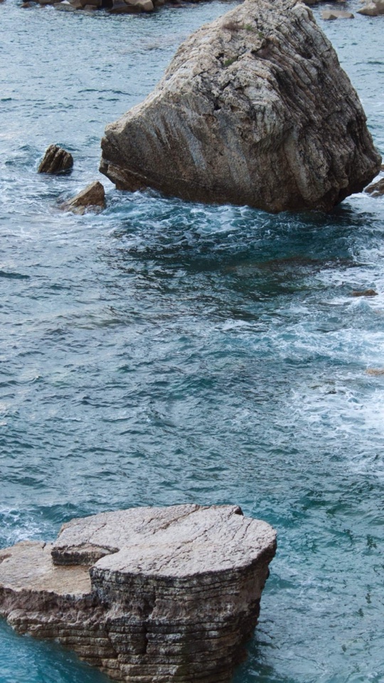 青い海に、大きな岩と平らな岩が浮かぶ荒々しい岩場の海岸をとらえた写真。