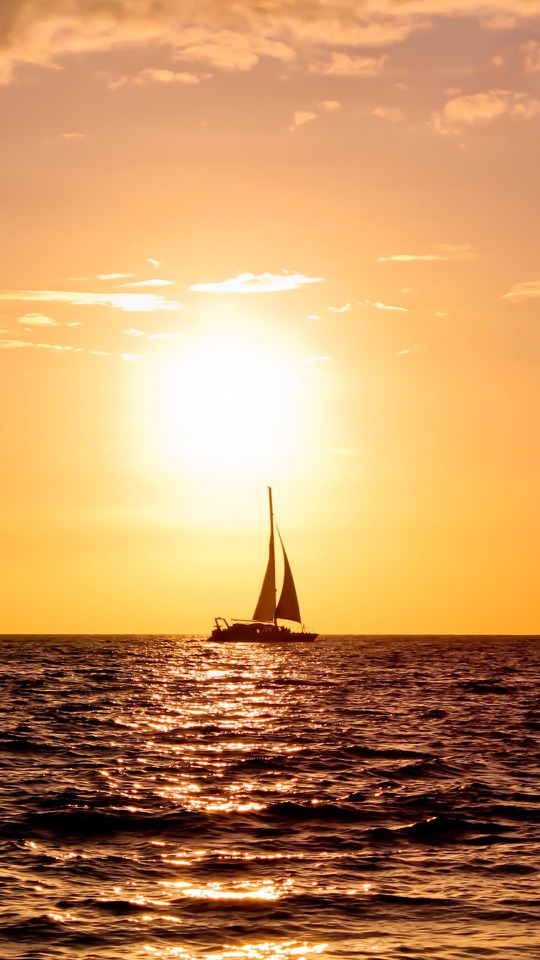 金色に染まる夕日の海に、帆船のシルエットが浮かぶ縦長の写真です。
