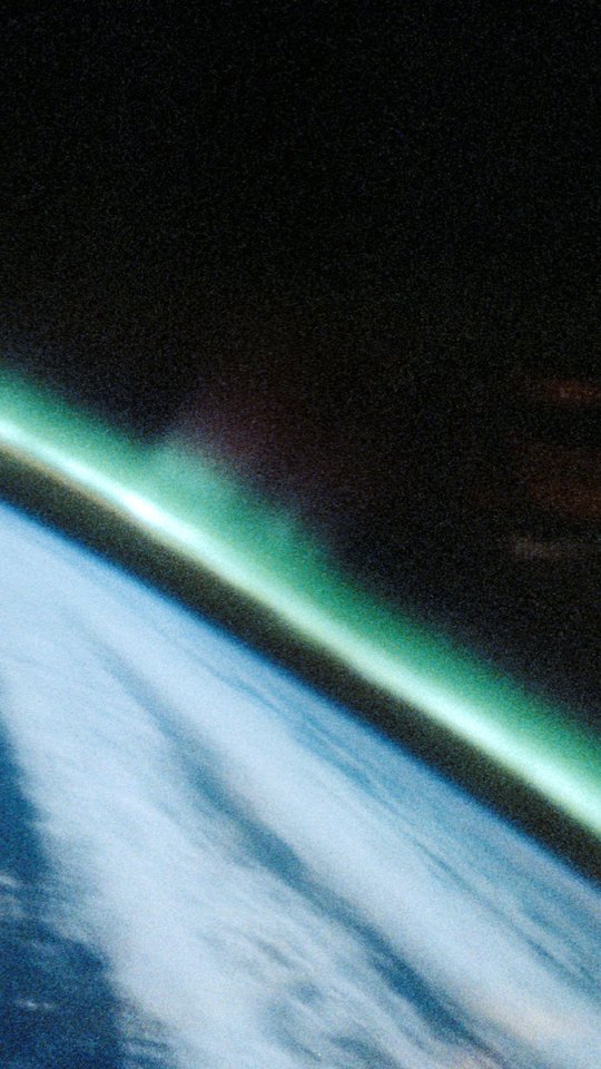 スペースシャトルから見た地球。白い雲に覆われた明るい地表の縁に、緑のオーロラが斜めに立つ。