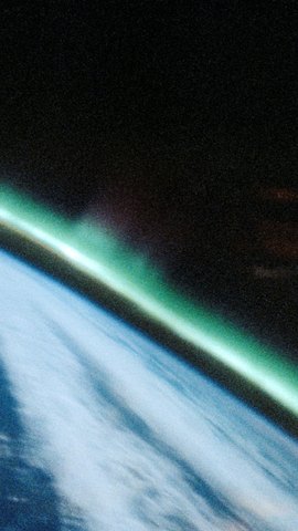 スペースシャトルから見た地球。白い雲に覆われた明るい地表の縁に、緑のオーロラが斜めに立つ。