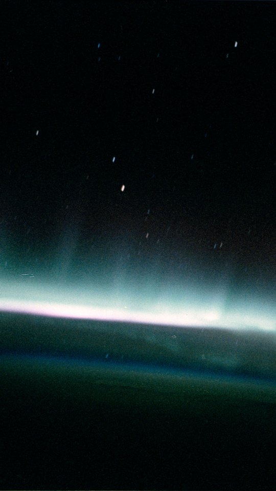 スペースシャトルから見たオーロラ。星の散る黒い空の下、地平線に沿って白とピンクの光の帯がにじむ。