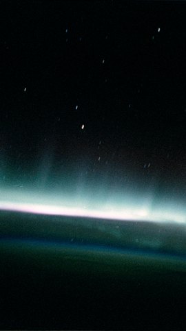 スペースシャトルから見たオーロラ。星の散る黒い空の下、地平線に沿って白とピンクの光の帯がにじむ。