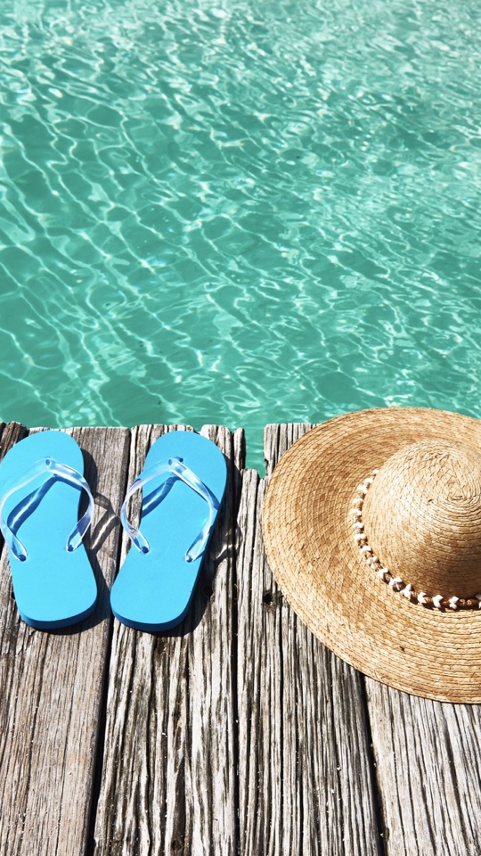 透き通ったプールの水辺に、青いビーチサンダルと麦わら帽子が置かれた夏の写真。