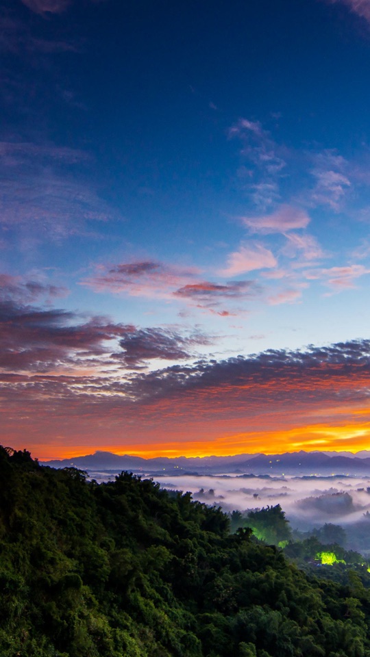 色鮮やかな夕焼けの空の下、霞のかかった緑の丘と谷が広がる風景写真。